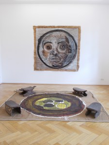 Wall painting: Portrait du President (2012), acrylic and tar on butcher's paper, 190 x 200 cm. Floor painting: Le Puits (2014), acrylic and tar on jute sacking, 245 x 280 cm. El Hadji Sy's artworks are displayed with four stools from Papua New Guinea that are part of the Weltkulturen Museum collection.
