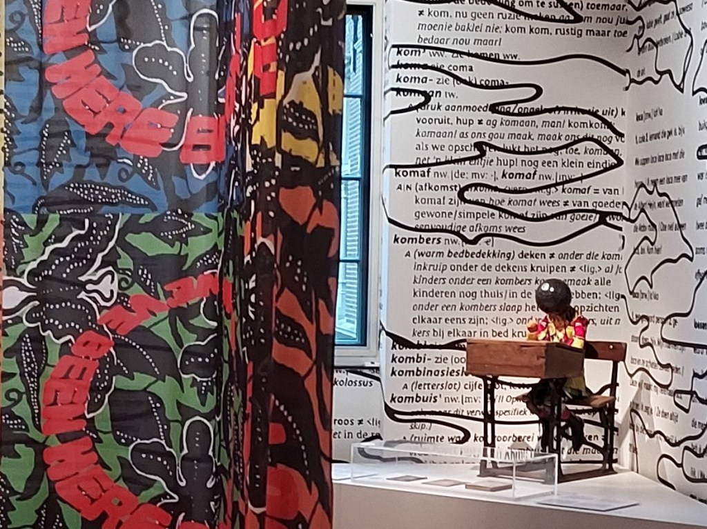 A museum-based art installation featuring a child-sized mannequin seated at a school desk, dressed in late-19th century clothing made from Dutch wax printed cotton fabric. A celestial globe replaces the head.