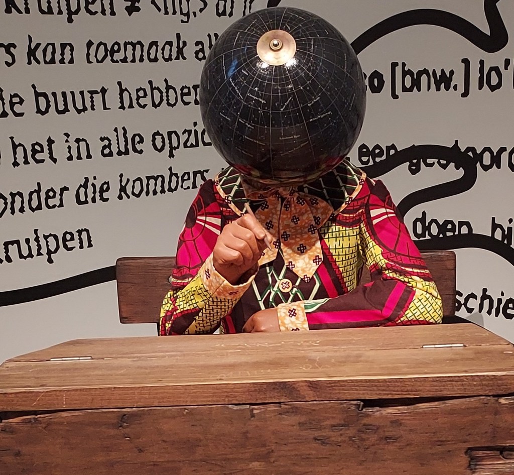 Mixed-media art installation featuring a child-sized mannequin seated at a wooden school desk, dressed in Victorian clothing made from patterned cotton fabrics, with a celestial globe replacing its head.