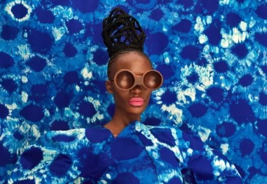 Photographic art installation featuring a young African woman dressed in an indigo tie-dyed fabric garment, with wooden eyewear obscuring her eyes, positioned against a backdrop made from the same fabric to create a camouflage effect.