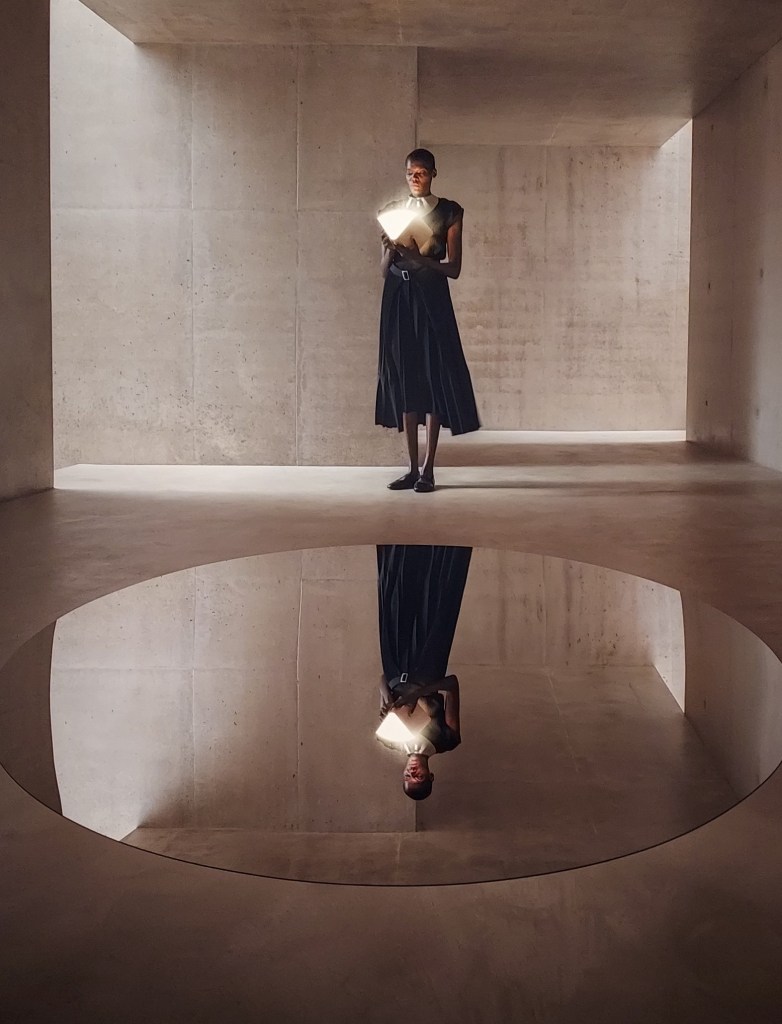 Woman holding an illuminated book, standing in front of a large, spherical, mirrored surface on the ground in front of her.