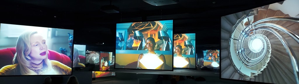 Multi-channel screening of a film-based art installation showing two female actors, each holding illuminated books, seated inside the post-modern architectural setting of Charles Jencks' Cosmic House in London. The third screen shows a spiral staircase.