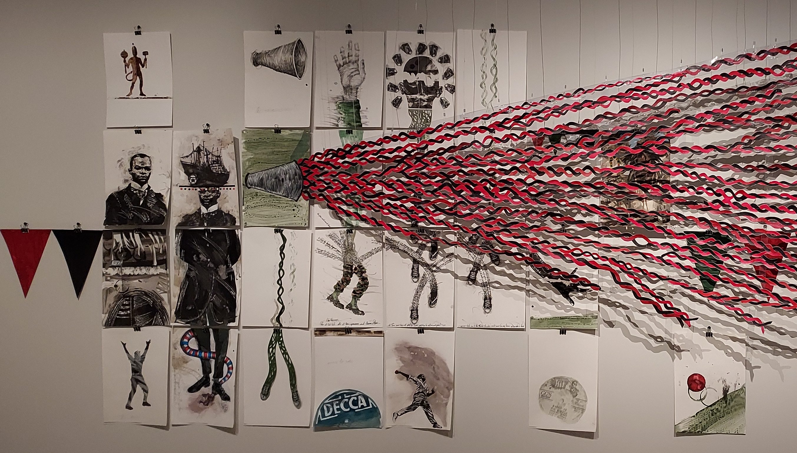 Photograph of a collage-based art installation by Christopher Cozier, featuring charcoal drawings and pencil sketches on c.25 small sheets of paper, collectively symbolising the history of Pan-Africanism. A group of intertwined red and black, ribbon-like strands of paper are also shown emanating from a drawing of a megaphone to represent the growth of the political and cultural movement in the form of a double helix.