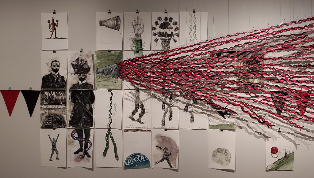 Photograph of a collage-based art installation by Christopher Cozier, featuring charcoal drawings and pencil sketches on c.25 small sheets of paper, collectively symbolising the history of Pan-Africanism. A group of intertwined red and black, ribbon-like strands of paper are also shown emanating from a drawing of a megaphone to represent the growth of the political and cultural movement in the form of a double helix.